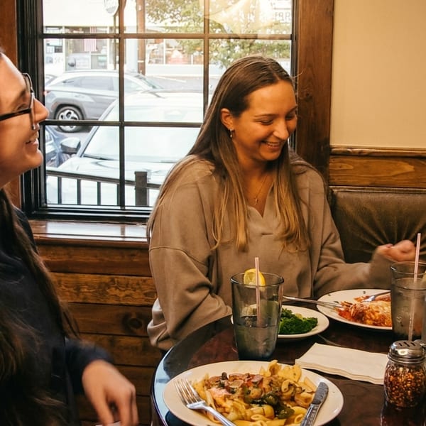 Two women smiling and eating pasta dishes at a restaurant table with drinks and spices.