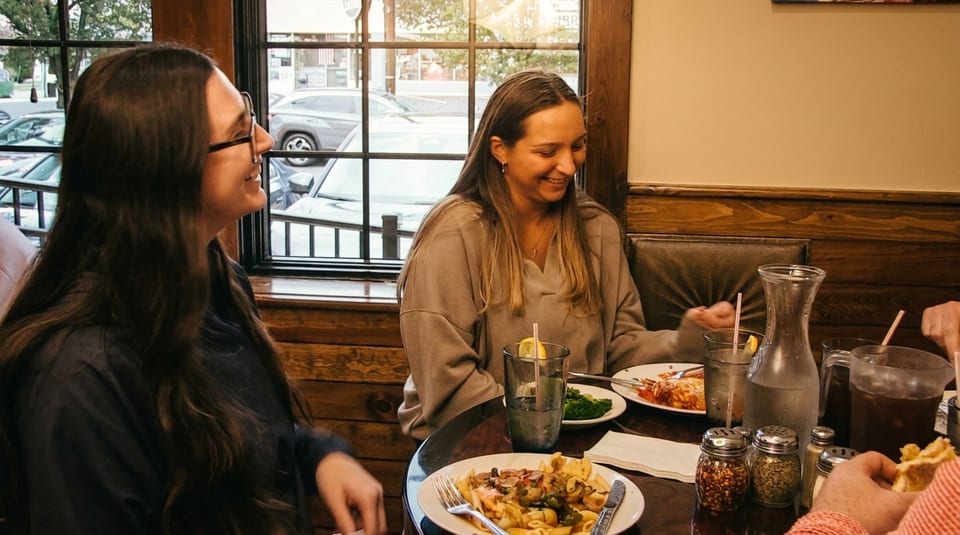 Two women smiling and eating pasta dishes at a restaurant table with drinks and spices.