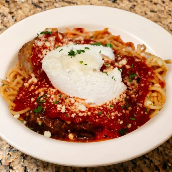 Spaghetti with red sauce, a large meatball, ricotta cheese, and parsley in a white bowl.