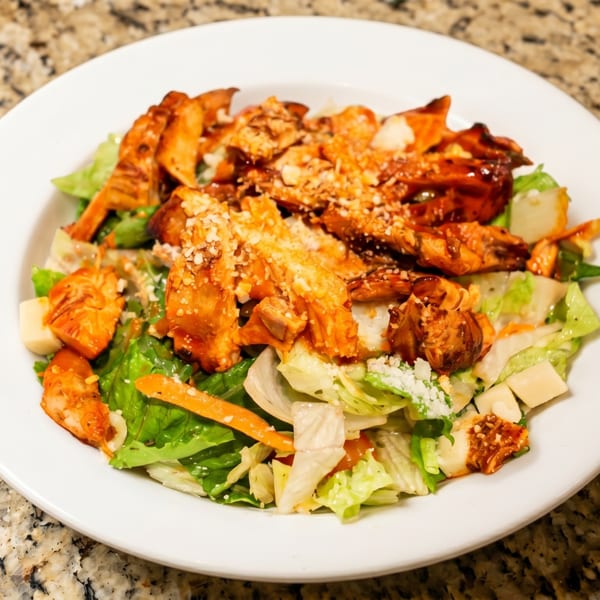 A white plate holds a salad with grilled chicken, lettuce, carrots, and parmesan cheese.