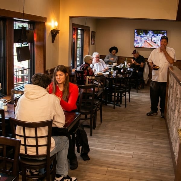 Diners seated at tables in a restaurant with a pizza display case and a chef statue.