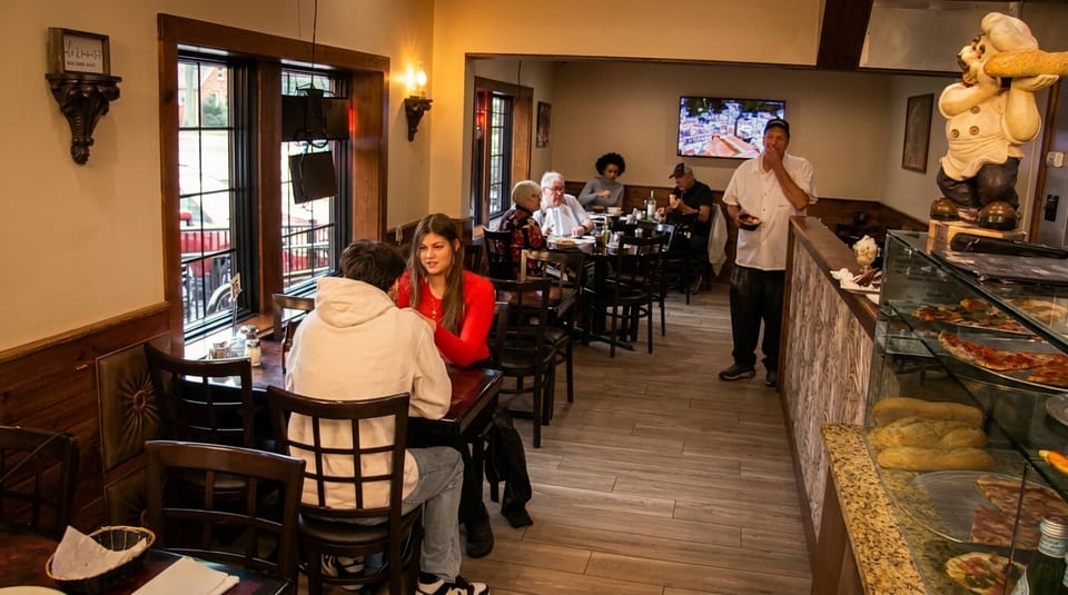 Diners seated at tables in a restaurant with a pizza display case and a chef statue.