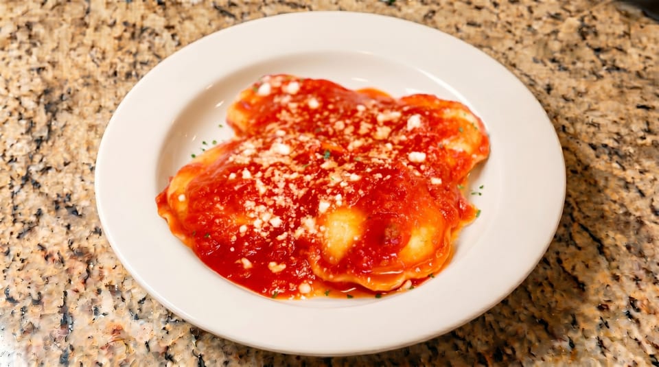 A white plate of ravioli covered in red sauce, sprinkled with white cheese and herbs.