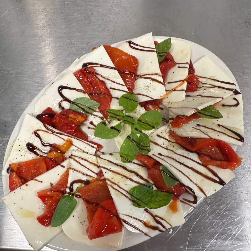 A white platter of mozzarella slices, roasted red peppers, basil, and balsamic glaze.