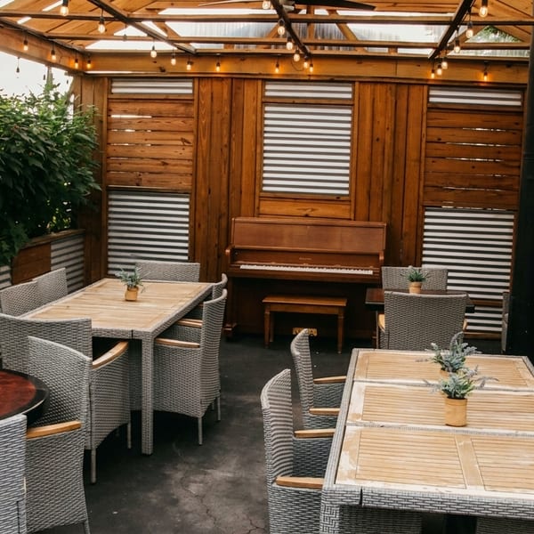 Outdoor patio dining area with wicker chairs, wooden tables, string lights, and a piano against a wood wall.