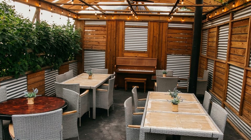 Outdoor patio dining area with wicker chairs, wooden tables, string lights, and a piano against a wood wall.